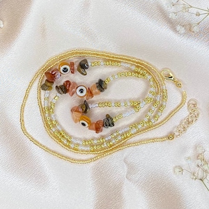 May include: A layered necklace with gold and white beads, featuring several evil eye charms and brown and orange stone beads.