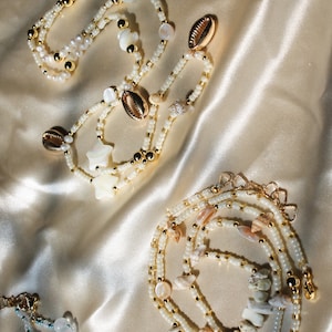 May include: Several necklaces with white beads, gold accents, and seashells are displayed on a satin surface. Some necklaces feature star-shaped and round shell accents. The jewelry has a beachy, coastal theme.
