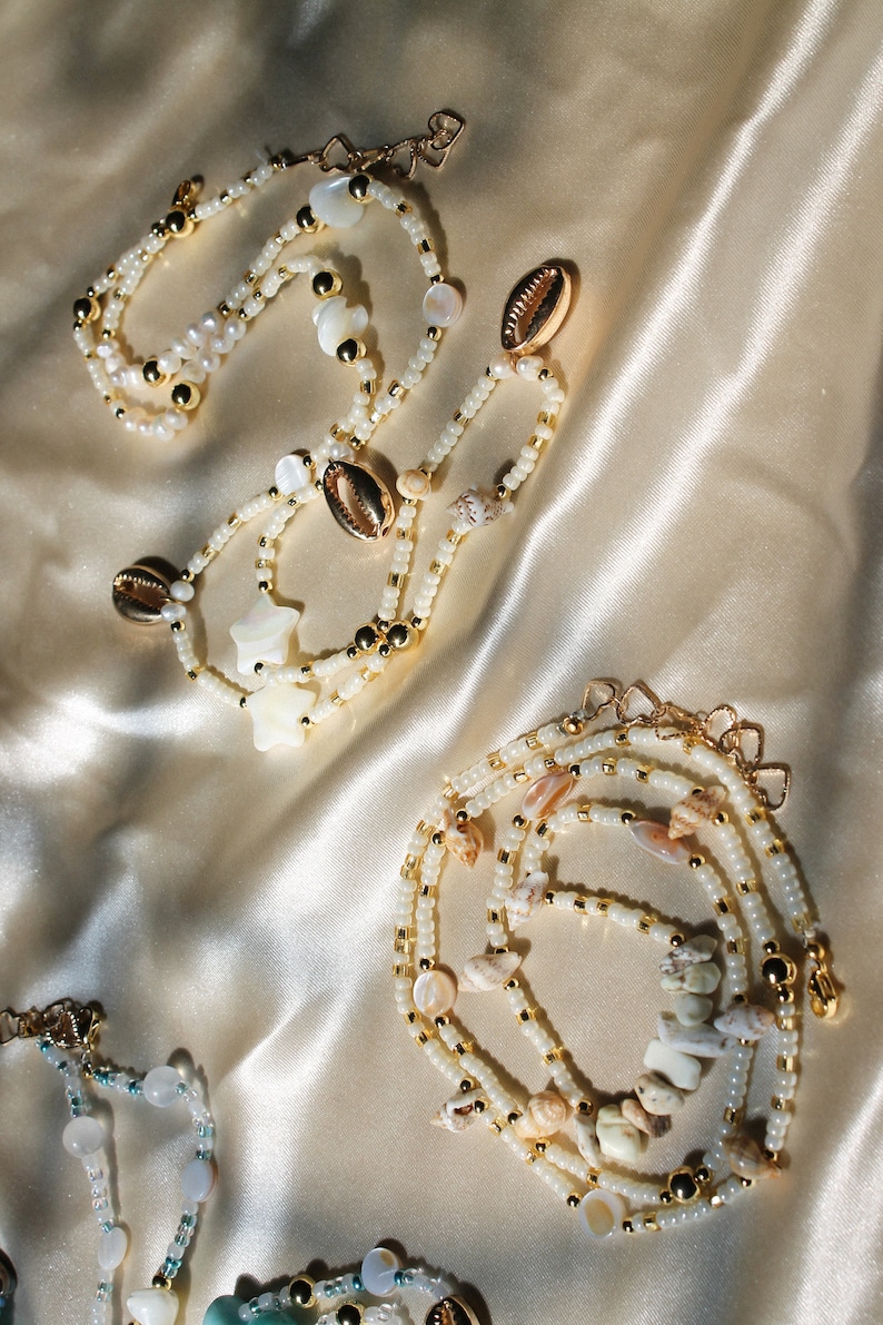 May include: Several necklaces with white beads, gold accents, and seashells are displayed on a satin surface. Some necklaces feature star-shaped and round shell accents. The jewelry has a beachy, coastal theme.