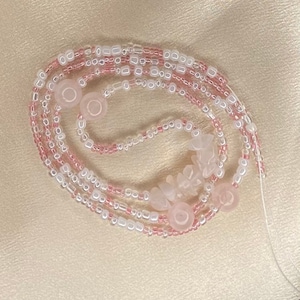 May include: A pink and white beaded necklace with rose quartz beads and charms. The necklace is made of multiple strands of beads and is tied together with a thin string.