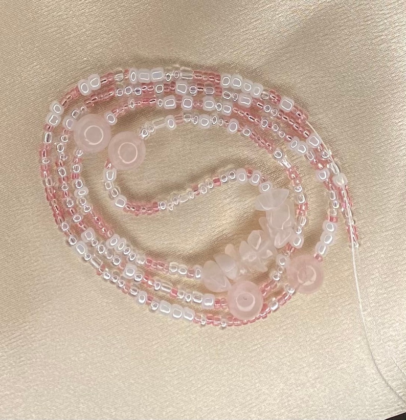 May include: A pink and white beaded necklace with rose quartz beads and charms. The necklace is made of multiple strands of beads and is tied together with a thin string.