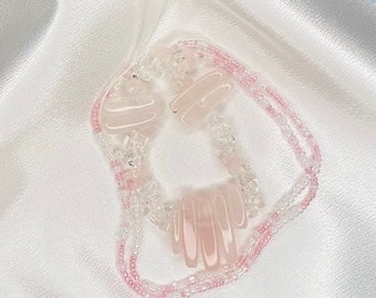 Puede incluir: Un collar de cuentas que presenta una combinación de cuentas de piedra rectangulares de color rosa pálido, cuentas de cristal transparente facetado y pequeñas cuentas de semillas rosas translúcidas dispuestas en un patrón circular.