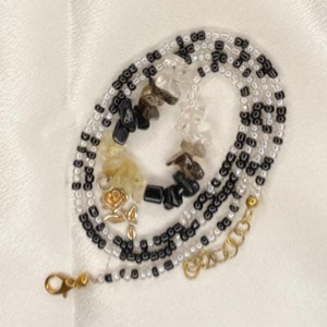 May include: A beaded necklace featuring black, white, and clear beads, with a gold-colored rose charm and a variety of natural stone chips. The necklace has a gold-colored clasp and extender chain.