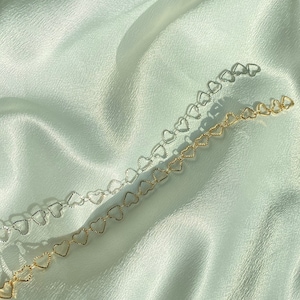 May include: Two necklaces with heart-shaped links, one silver and one gold. They are displayed on a white, silky fabric. The heart-shaped design adds a touch of charm. The necklaces are delicate and create a soft, romantic aesthetic.