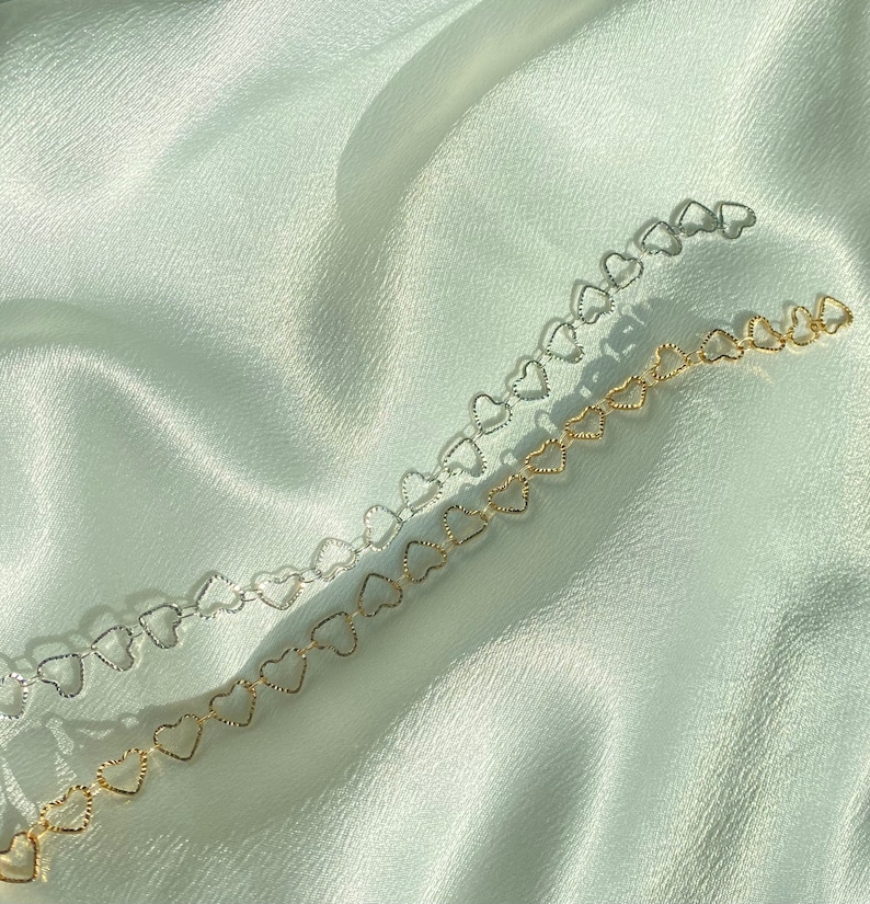 May include: Two necklaces with heart-shaped links, one silver and one gold. They are displayed on a white, silky fabric. The heart-shaped design adds a touch of charm. The necklaces are delicate and create a soft, romantic aesthetic.