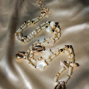 Peut inclure: Un collier à plusieurs rangs avec des perles blanches, des perles dorées et des breloques en forme de coquillages. Le collier présente des breloques en forme d'étoile et de coquillages.