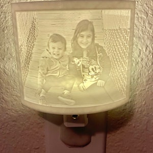 May include: A white ceramic nightlight with a 3D image of two children. The children are smiling and looking at the camera. The nightlight is plugged into an electrical outlet.