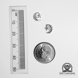 Può includere: Un paio di orecchini d'argento con un design a forma di luna crescente con una stella all'interno. Gli orecchini sono mostrati accanto a una moneta da dieci centesimi di dollaro americano per il confronto delle dimensioni. East Havana Jewelry.