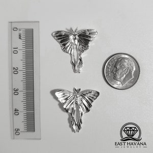 May include: Two silver-colored moth pendants with detailed wing patterns are displayed. A ruler shows measurements in millimeters, and a dime is included for size comparison. The pendants are on a white surface. The East Havana Jewelry logo is visible.