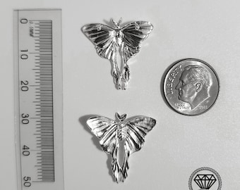Può includere: Due ciondoli a forma di falena color argento con dettagliate fantasie alari sono esposti. Un righello mostra le misure in millimetri e una moneta è inclusa per il confronto delle dimensioni. I ciondoli sono su una superficie bianca. Il logo East Havana Jewelry è visibile.