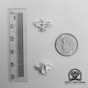 May include: Two silver bee charms, one slightly larger than the other, are shown next to a US dime for size comparison. The charms are detailed with raised lines and a textured surface. The smaller charm is about the size of a dime. The larger charm is slightly bigger than a dime. The charms are on a white background.  East Havana Jewelry is printed at the bottom of the image.