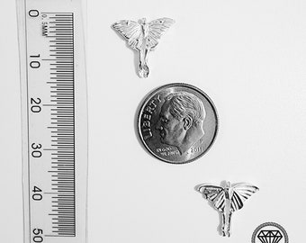 Può includere: Due ciondoli a forma di falena color argento e una moneta da dieci centesimi americana sono esposti su una superficie bianca. Un righello con segni millimetrici è posizionato a sinistra. I ciondoli hanno ali dettagliate.