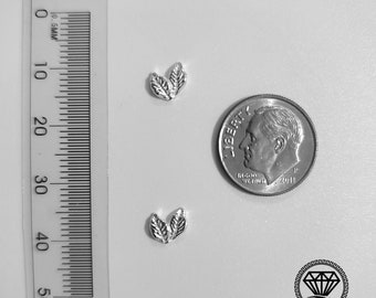 Può includere: Due ciondoli a forma di foglia in argento, ognuno con due foglie, sono mostrati accanto a una moneta da dieci centesimi di dollaro americano per il confronto delle dimensioni. I ciondoli sono piccoli e delicati, perfetti per aggiungere un tocco di natura ai gioielli.