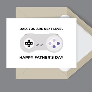 May include: A Father's Day card featuring a grey video game controller. The card has the text "DAD, YOU ARE NEXT LEVEL" and "HAPPY FATHER'S DAY" in black. It sits inside a beige envelope.