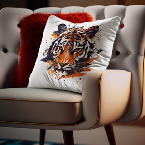K&ouml;nnte beinhalten: Ein wei&szlig;es Kissen mit einem farbenfrohen, abstrakten Tigergesichtsdesign. Das Kissen liegt auf einem hellgrauen Sessel mit einem roten Kissen.