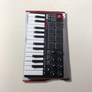 Pode incluir: Um teclado Akai MPK mini preto e branco com 25 teclas, 8 pads de bateria e 8 botões. O teclado está em uma caixa vermelha e branca com o logotipo da Akai.