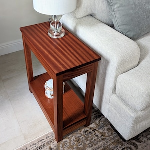 May include: A wooden end table with a single shelf. The table is made of rich mahogany wood with a natural grain pattern. A small decorative box sits on the shelf. A lamp with a white shade sits on the top shelf.