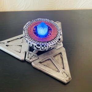 May include: A silver and brown 3D printed model of a sci-fi device with a glowing blue crystal in the center.