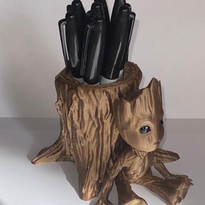Puede incluir: Una figurita Groot impresa en 3D de color marrón que sostiene un porta lápices con forma de tronco de árbol. El porta lápices está lleno de bolígrafos negros.