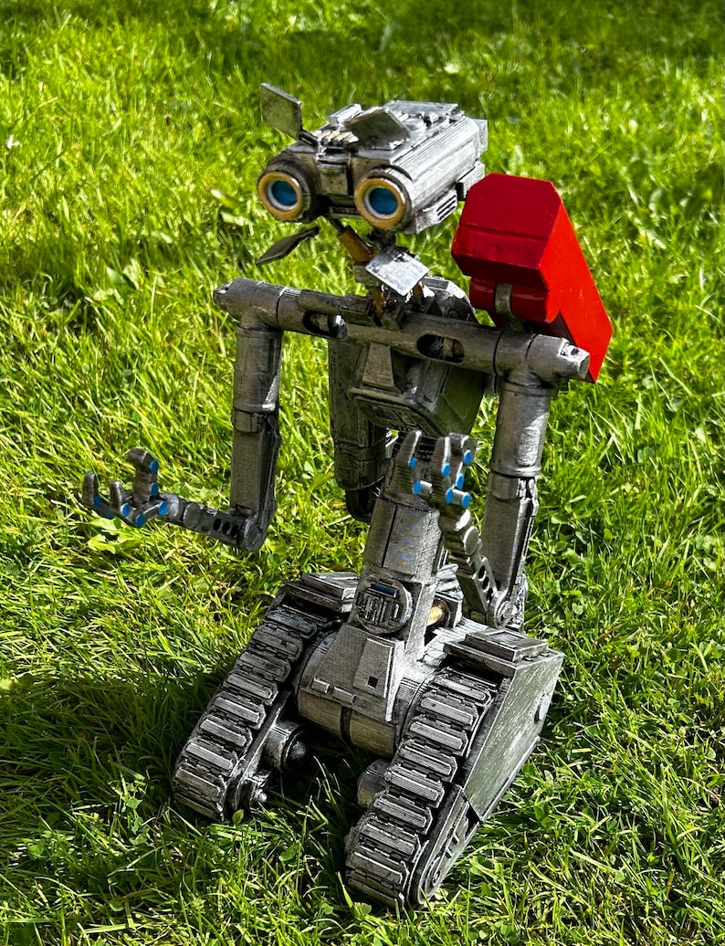 May include: A silver robot with blue eyes and a red backpack is kneeling on green grass. The robot has a tracked base and appears to be made of metal.