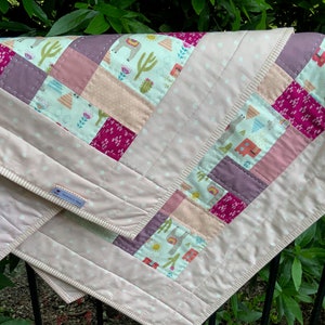 May include: A pink and white baby quilt with a patchwork design featuring llamas, cacti, and geometric shapes. The quilt has a white polka dot backing and a pink and white striped border.