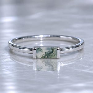Può includere: Un anello d'argento con una pietra di agata muschiata rettangolare. La pietra presenta un motivo naturale verde e bianco. L'anello ha una fascia sottile e un design minimalista. L'anello è mostrato su una superficie riflettente.