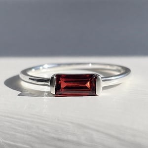 Puede incluir: Un anillo de plata con una piedra preciosa de granate rojo intenso, tallada en forma de baguette rectangular y engastada horizontalmente en un bisel de metal liso. La banda es fina, pulida y circular, con un diseño limpio y una piedra central.