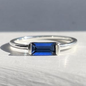 May include: A silver ring featuring a single baguette-cut deep blue gemstone set horizontally in the centre. The band is smooth and polished, with the stone held securely by two metal prongs on either side, creating a clean and simple design.