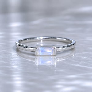 Puede incluir: Un anillo de plata con una piedra preciosa rectangular e iridiscente. El anillo tiene una banda fina y pulida, y la piedra preciosa está engastada en un bisel rectangular. La piedra preciosa muestra una gama de colores, incluyendo azul y blanco.