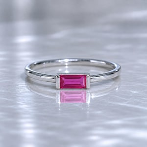 May include: A silver ring featuring a rectangular, vivid pink gemstone. The ring has a slender, polished band, and the gemstone is set in a minimalist, contemporary design. The ring is shown on a reflective surface.