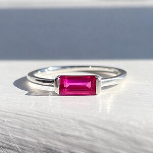 May include: A silver ring featuring a single rectangular baguette-cut vibrant pink gemstone set horizontally in a smooth metal band. The stone is held in place by two silver prongs on either side, creating a clean and simple design for the finger.