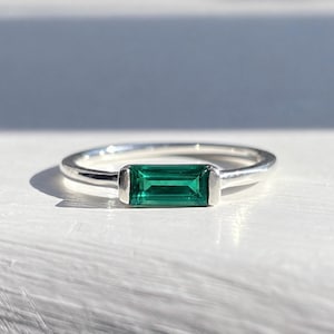 Peut inclure: Une bague en argent ornée d'une pierre précieuse rectangulaire taille baguette d'un vert émeraude, sertie horizontalement dans une monture en métal lisse. L'anneau est fin et poli, présentant un design épuré avec la pierre verte.
