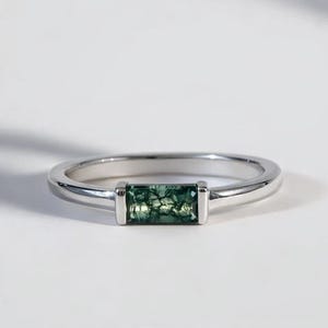 Puede incluir: Un anillo de plata con una gema rectangular verde musgo. El anillo tiene una banda simple y un engaste de bisel. La gema tiene un patrón natural, y el anillo está sobre un fondo blanco. Una joya elegante para cualquier ocasión.