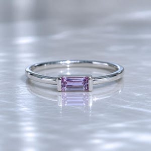 Puede incluir: Un anillo de plata con una piedra preciosa rectangular de color púrpura claro. La piedra preciosa está engastada horizontalmente en el centro del anillo. La banda es delgada y pulida, reflejando la luz. El anillo se muestra sobre una superficie clara y reflectante.