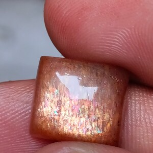 Op de afbeelding: Een vierkante, iriserende sunstone cabochon met een roodbruine basis en een schitterend, regenboogachtig effect. De steen is ongeveer 2,5 cm groot.