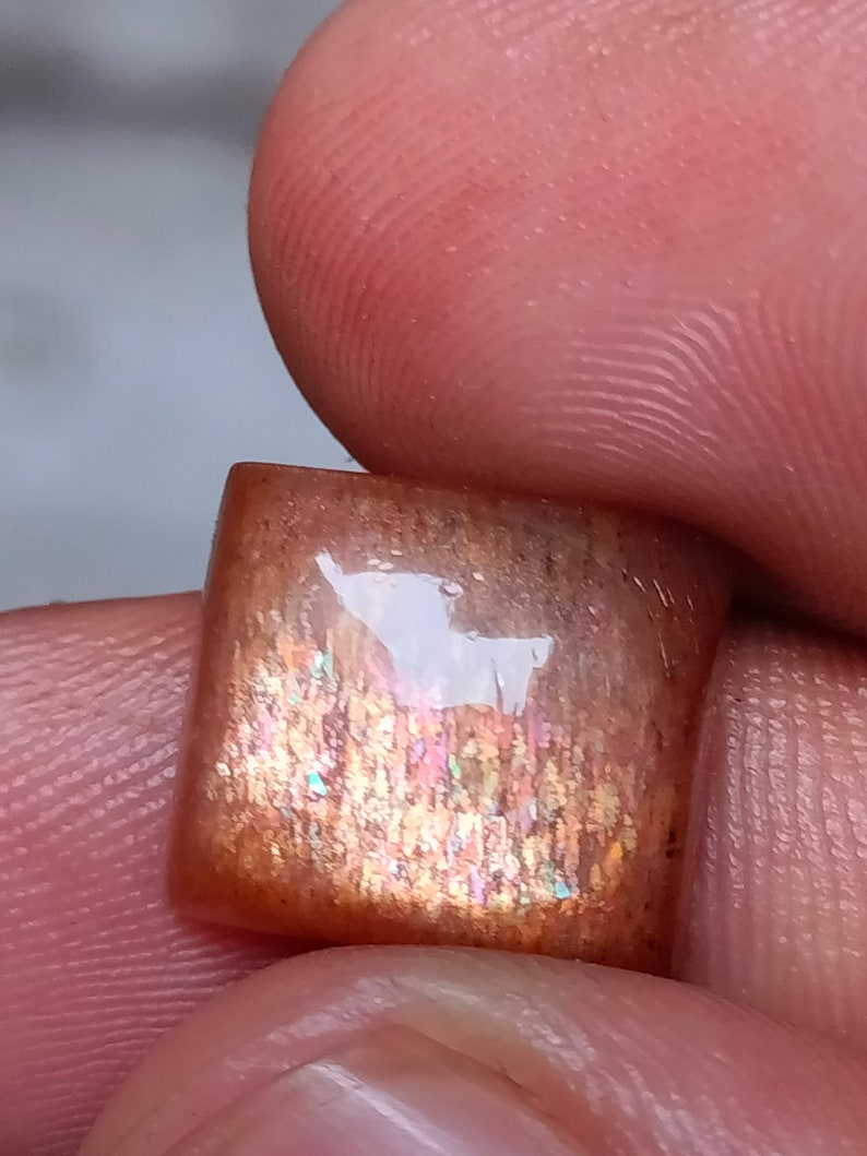 Op de afbeelding: Een vierkante, iriserende sunstone cabochon met een roodbruine basis en een schitterend, regenboogachtig effect. De steen is ongeveer 2,5 cm groot.