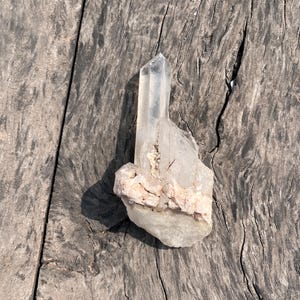 May include: A clear quartz crystal cluster with a pointed tip and a rough, light-colored base. The crystal is transparent, allowing light to pass through, and sits on a weathered, gray wooden surface. The quartz has natural inclusions.
