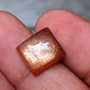 Op de afbeelding: Een vierkante, iriserende sunstone cabochon met een roodbruine kleur en een glinsterende, metallic glans.