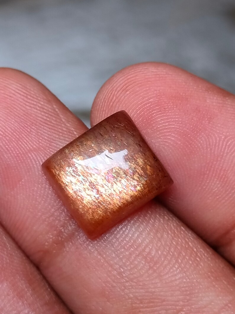 Op de afbeelding: Een vierkante, iriserende sunstone cabochon met een roodbruine kleur en een glinsterende, metallic glans.