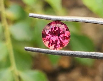 Puede incluir: Una piedra preciosa redonda y facetada de un tono rojo rosado vibrante, sostenida por unas pinzas de metal plateado. La piedra muestra múltiples facetas geométricas que reflejan la luz, creando un aspecto brillante y centelleante.
