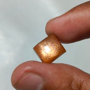 Op de afbeelding: Een vierkante, gepolijste sunstone edelsteen met een roodbruine kleur en een glinsterende, iriserende afwerking.