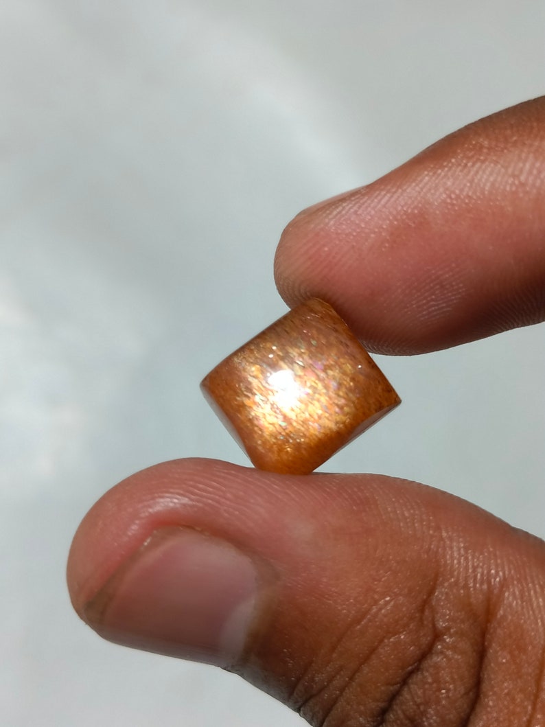 Op de afbeelding: Een vierkante, gepolijste sunstone edelsteen met een roodbruine kleur en een glinsterende, iriserende afwerking.