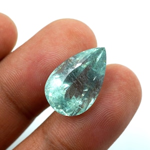 May include: A pear-shaped, faceted gemstone with a light teal color. The gem is held between two fingers against a white background. The gem has a polished surface with internal inclusions.