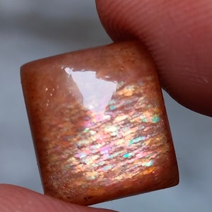 Op de afbeelding: Een rechthoekige, iriserende sunstone cabochon met een bruine basis en een regenboog van kleuren die erin schitteren.