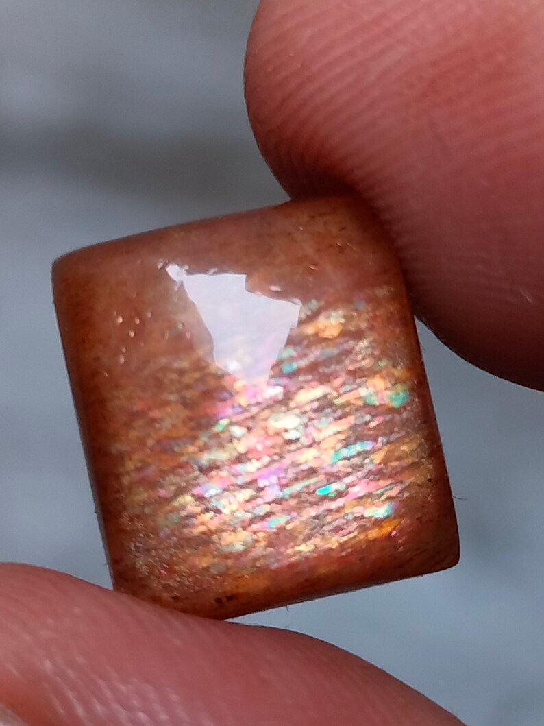 Op de afbeelding: Een rechthoekige, iriserende sunstone cabochon met een bruine basis en een regenboog van kleuren die erin schitteren.