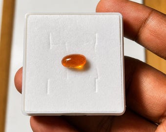 Puede incluir: Una pequeña piedra preciosa de color naranja translúcido con forma ovalada y superficie lisa y pulida. La piedra está centrada sobre un inserto de espuma blanca cuadrado dentro de una caja de presentación de plástico blanco brillante.