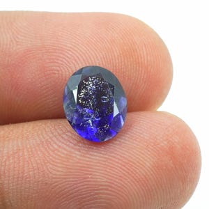 Puede incluir: Una gema ovalada facetada, de color azul profundo con inclusiones brillantes. La gema se sostiene entre los dedos, mostrando su superficie pulida y brillo interno. Las facetas de la piedra capturan la luz, creando un efecto brillante.