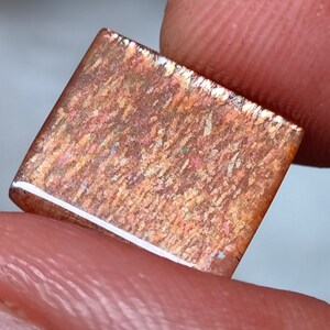 Op de afbeelding: Een rechthoekige, iriserende edelsteen met een bruine, oranje en gouden kleur. De edelsteen heeft een getextureerd oppervlak en een witte rand.