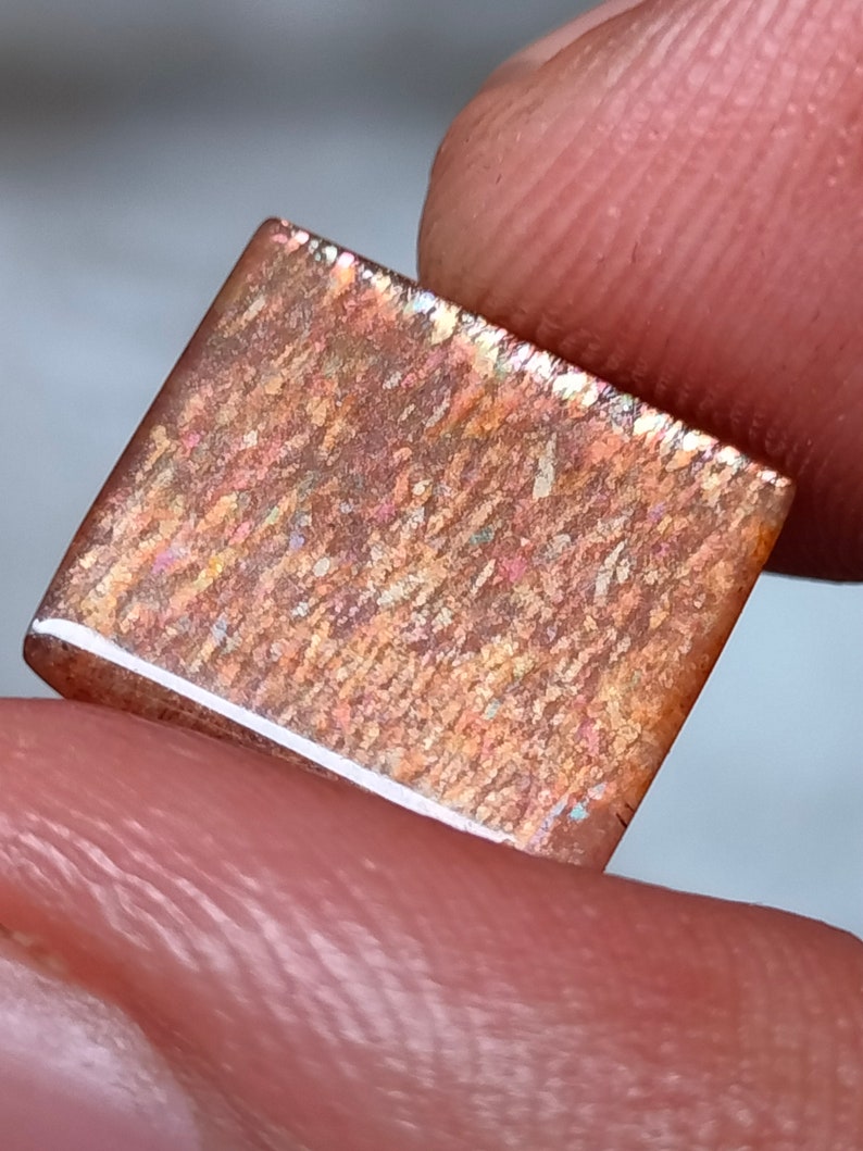 Op de afbeelding: Een rechthoekige, iriserende edelsteen met een bruine, oranje en gouden kleur. De edelsteen heeft een getextureerd oppervlak en een witte rand.
