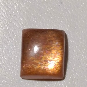 Op de afbeelding: Een vierkante, iriserende sunstone cabochon met een roodbruine kleur en een glinsterende, glimmende afwerking.